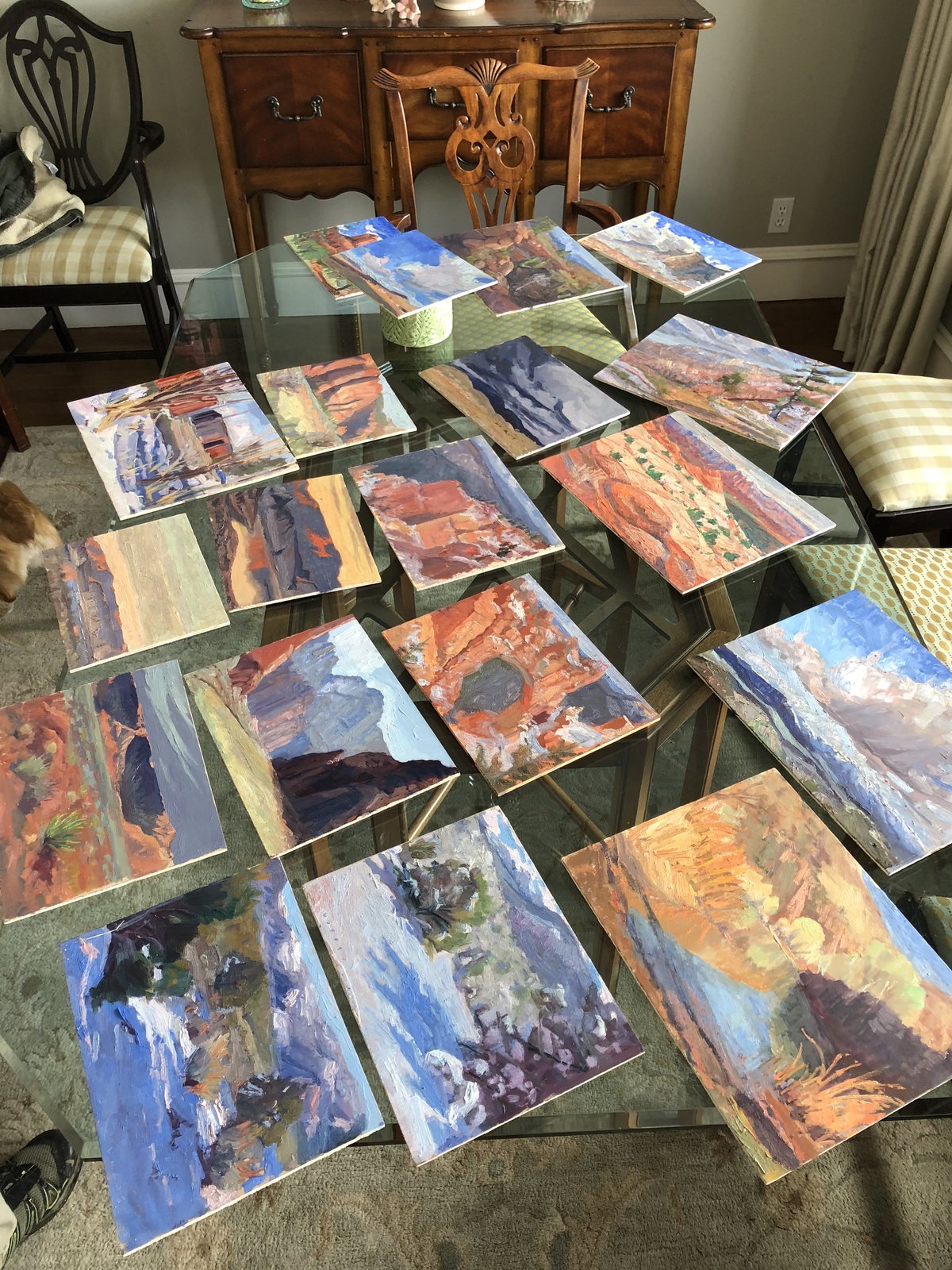 About twenty small oil studies laid out on a glass table, desert reds, snow and sky