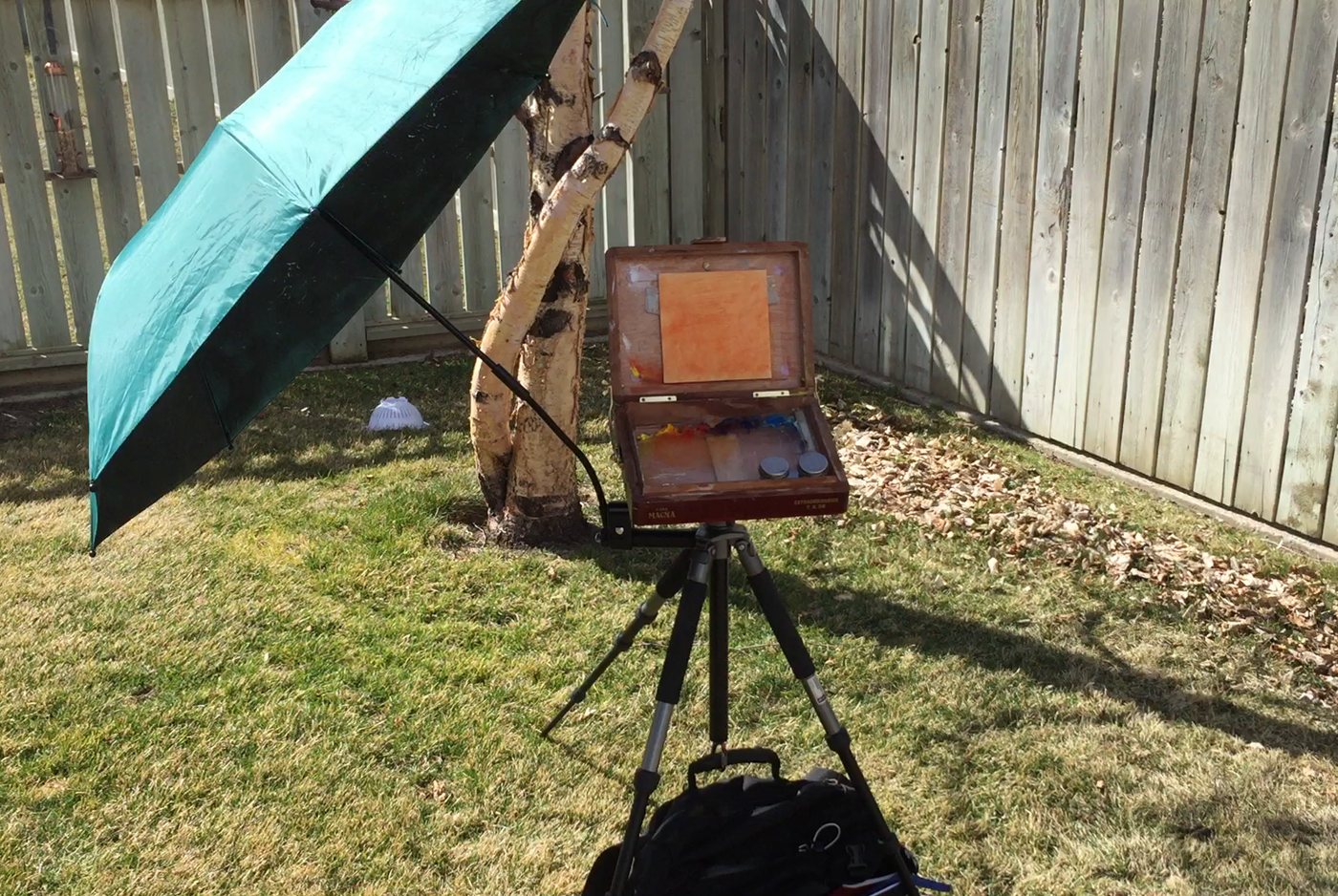 The red easel on a tripod under a green umbrella, a bag hanging below