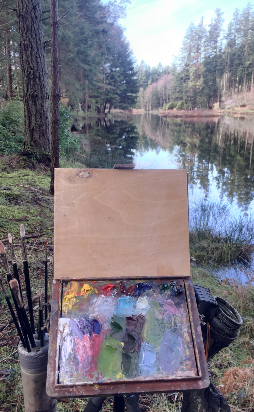 The first box with a full palette and the brush holder beside a still forest lake