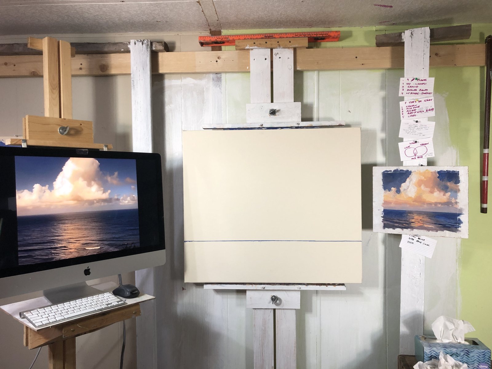 The studio easel with a blank toned panel, the small cloud sketch pinned beside it, the photo on a monitor